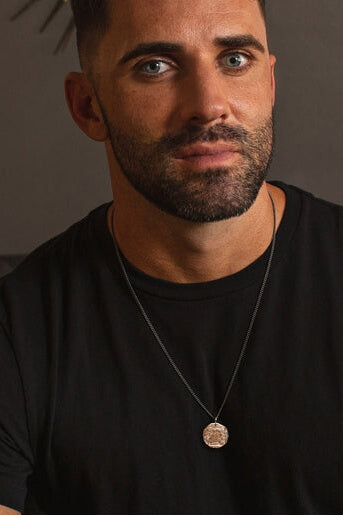Man wearing a black t-shirt and necklace with a pendant against a neutral background