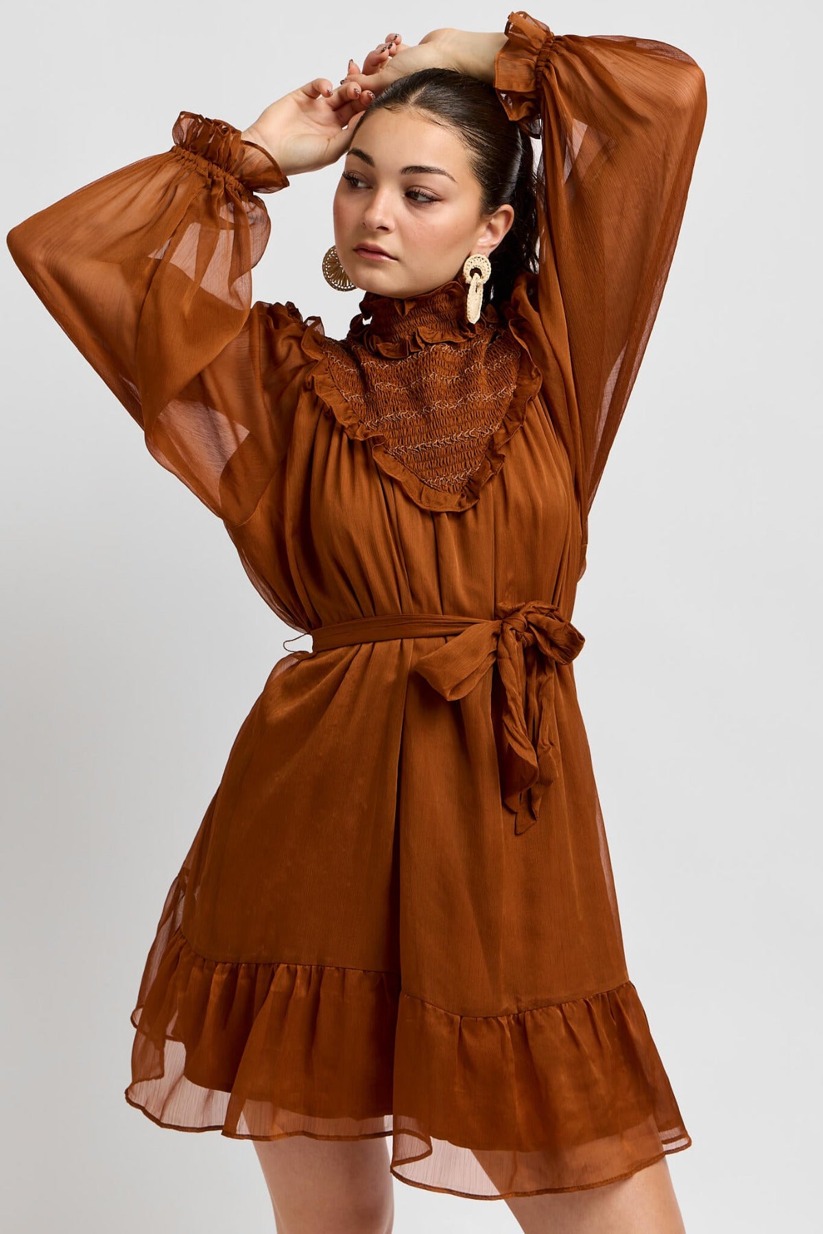 Woman wearing a brown dress with sheer sleeves and ruffled details on a white background